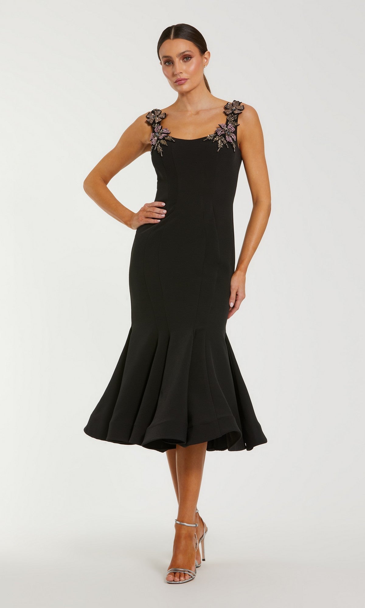 A woman stands against a plain background wearing the black Mac Duggal 11948 Midi Wedding Guest Dress with fitted silhouette, sleeveless design, 3D floral-embellished straps, flared hem, and silver high-heeled sandals.